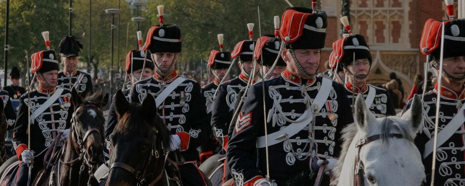A regiment of historical cavalry soldiers on horseback wearing bespoke dark ceremonial uniforms with white braiding and shako hats.